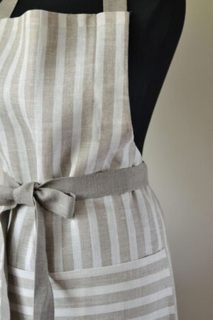 Striped linen apron with adjustable belt and pocket. Stipes goes along the apron natural linen color with off white. Belt made from natural linen. And the apron's pocket made from the stipes goes across the apron.