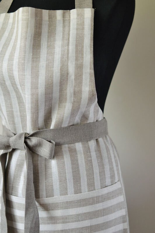 Striped linen apron with adjustable belt and pocket. Stipes goes along the apron natural linen color with off white. Belt made from natural linen. And the apron's pocket made from the stipes goes across the apron.