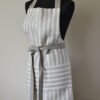 Striped linen apron with adjustable belt and pocket. Stipes goes along the apron natural linen color with off white. Belt made from natural linen. And the apron's pocket made from the stipes goes across the apron.