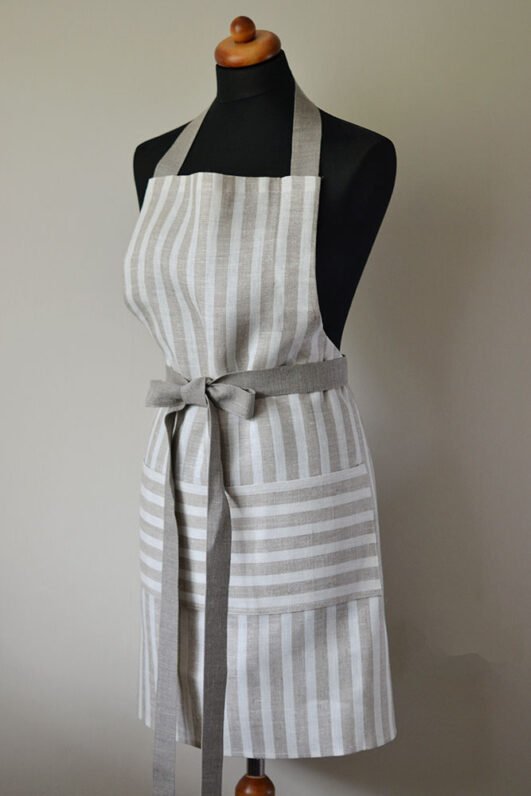 Striped linen apron with adjustable belt and pocket. Stipes goes along the apron natural linen color with off white. Belt made from natural linen. And the apron's pocket made from the stipes goes across the apron.
