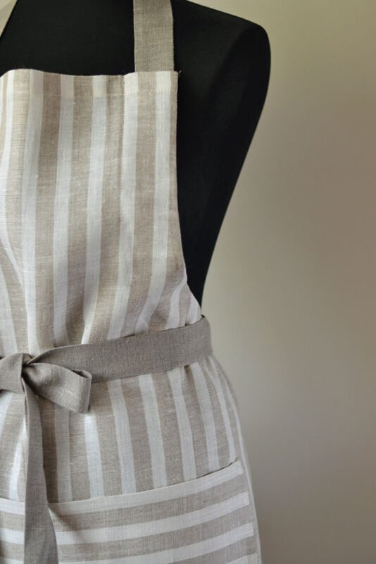 Striped linen apron with adjustable belt and pocket. Stipes goes along the apron natural linen color with off white. Belt made from natural linen. And the apron's pocket made from the stipes goes across the apron.
